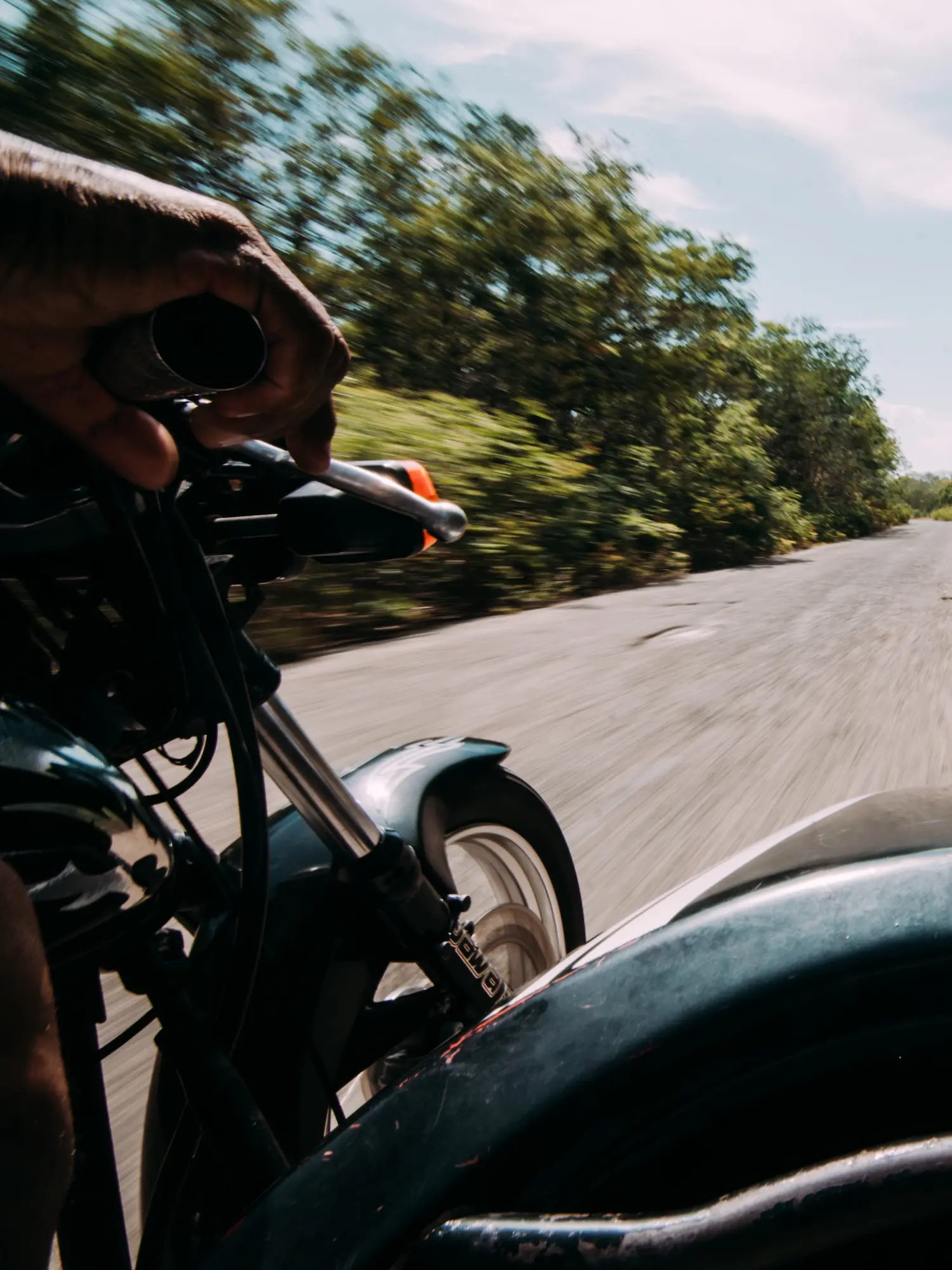 A point-of-view shot shows a person riding a black motorcycle down a rural road.
