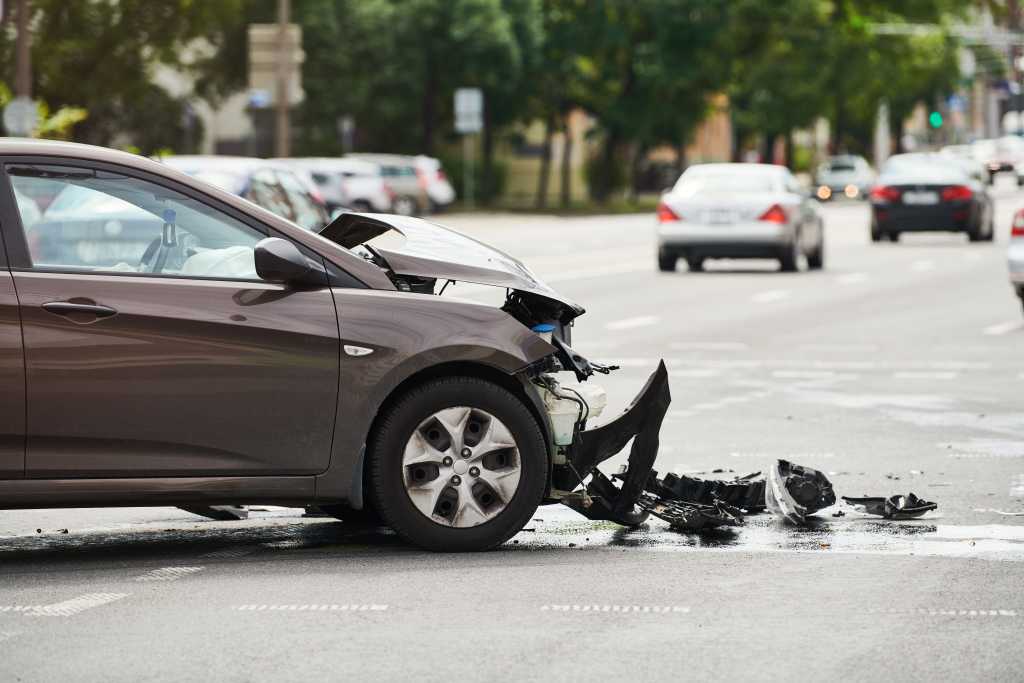 A brown sedan with severe front-end damage stopped at an intersection in kansas city, with the hood crumpled, the bumper detached, and debris scattered across the road. Traffic and tree-lined streets are visible in the background.