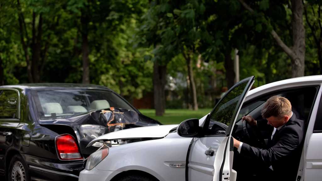 A man in a dark suit steps out of a white car that has been involved in a rear-end collision with a black sedan. Both vehicles show significant front and rear damage. The scene takes place on a tree-lined street on a sunny day.